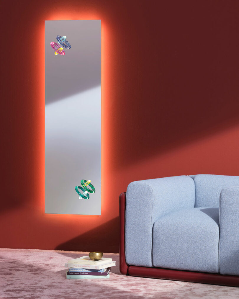 A tall, rectangular mirror with an orange backlight is mounted on a red wall above a stack of books, next to a light blue armchair on a pink rug.