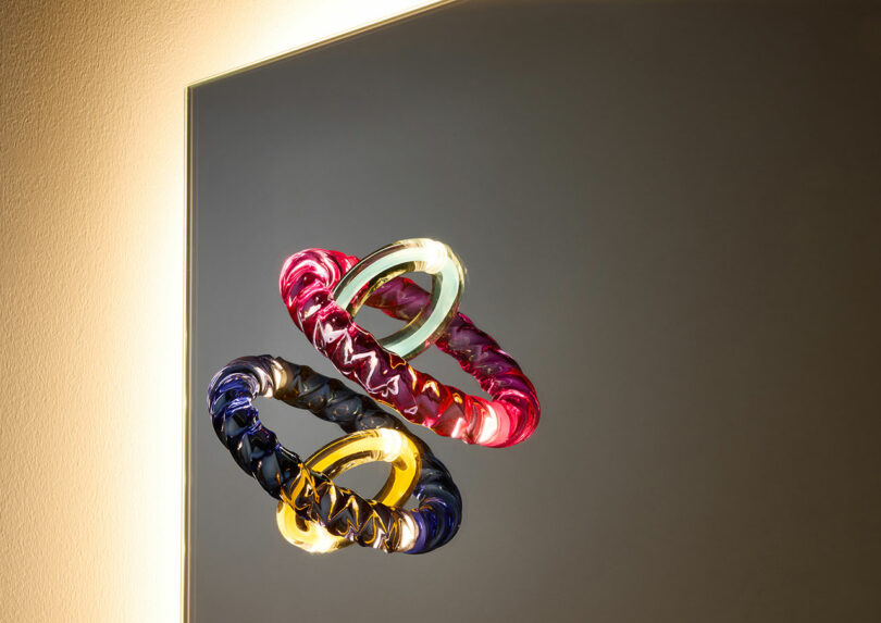 Two illuminated, intertwined rings—one pink and one blue with yellow accents—are suspended in front of a reflective surface, casting soft light on the surroundings.