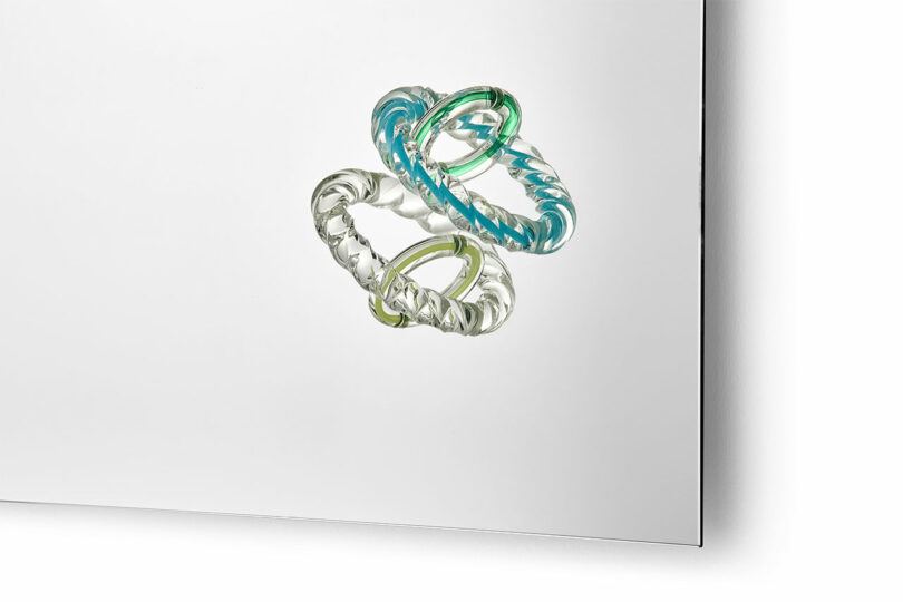 A clear glass surface with three interlocking, twisted glass rings—blue, green, and transparent—mounted near one edge in the signature Hooptical style of Bethan Laura Wood.