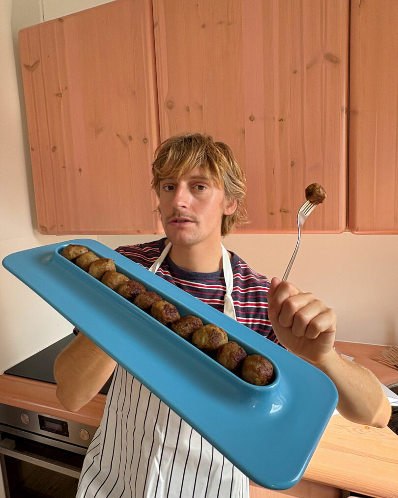 The person in the striped shirt and the aprons keep a blue tray with meatballs and shows one meatball on a fork in the kitchen.