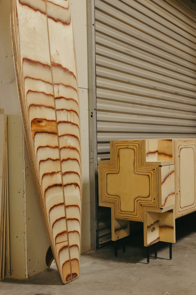 Blätter von Sperrholz mit sichtbarem Holzkorn lehnen sich gegen die Wand, neben zwei Holzstrukturen mit einem Kreuzschnitt an schwarzen Metallbeinen-der Szene eines Workshops mit einem Hauch eines von Kelly Wearstler inspirierten Stils heißt.