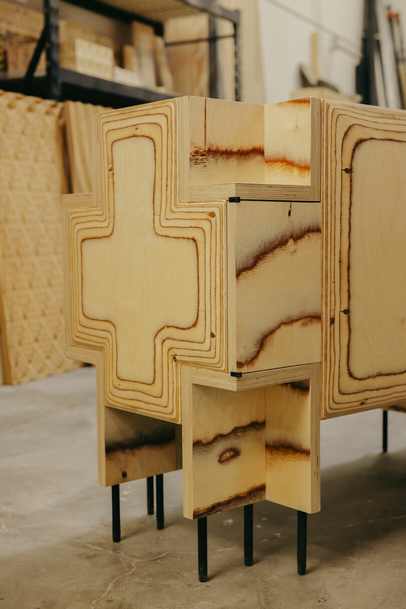 Ein Holzschrank mit einem geometrischen Design in Form eines Kreuz- und schwarzen Metallbeine, die an einen mutigen Stil von Kelly Wearstler erinnern, steht in einem Workshop mit Werkzeugen und Holz im Hintergrund.