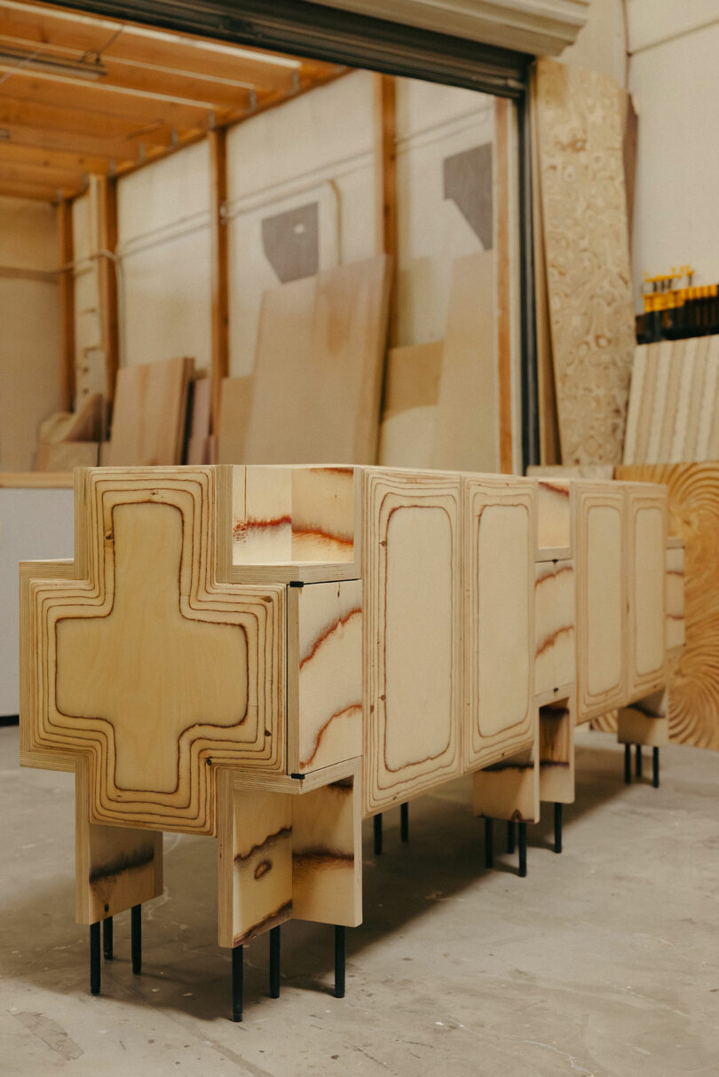 Ein Holzschrank mit Kreuzenden und geschichtter Sperrholzdesign, inspiriert von Kelly Wearstler, steht in einer Holzverarbeitungsgeschäft an schwarzen Metallbeinen.