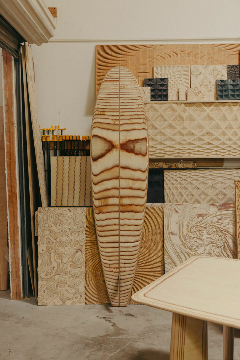 Die hölzerne Surfplatte mit einem symmetrischen Kornmuster steht aufrecht gegen die Wand, umgeben von verschiedenen Sperrholzblättern, die komplexe Muster im Workshop haben, die an den mutigen Stil von Kelly Wearstler erinnern.