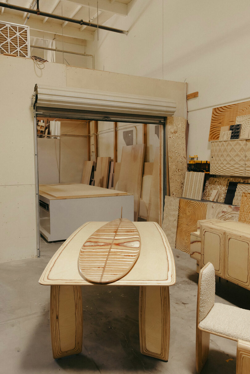 Surfbrett mit Getreideholzmuster befindet sich auf dem entsprechenden Holztisch im Werkstatt mit Sperrholzblättern und unvollendeten Möbeln und spiegelt den raffinierten Stil von Kelly Wearstler wider.