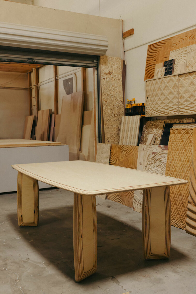 Ein hölzerner Tisch mit dicken gebogenen Beinen steht in einer Werkstatt, umgeben von Blättern aus gemusterten Holz- und Holzwerkzeugen, die an mutige und einzigartige Muster von Kelly Wearstler erinnern.