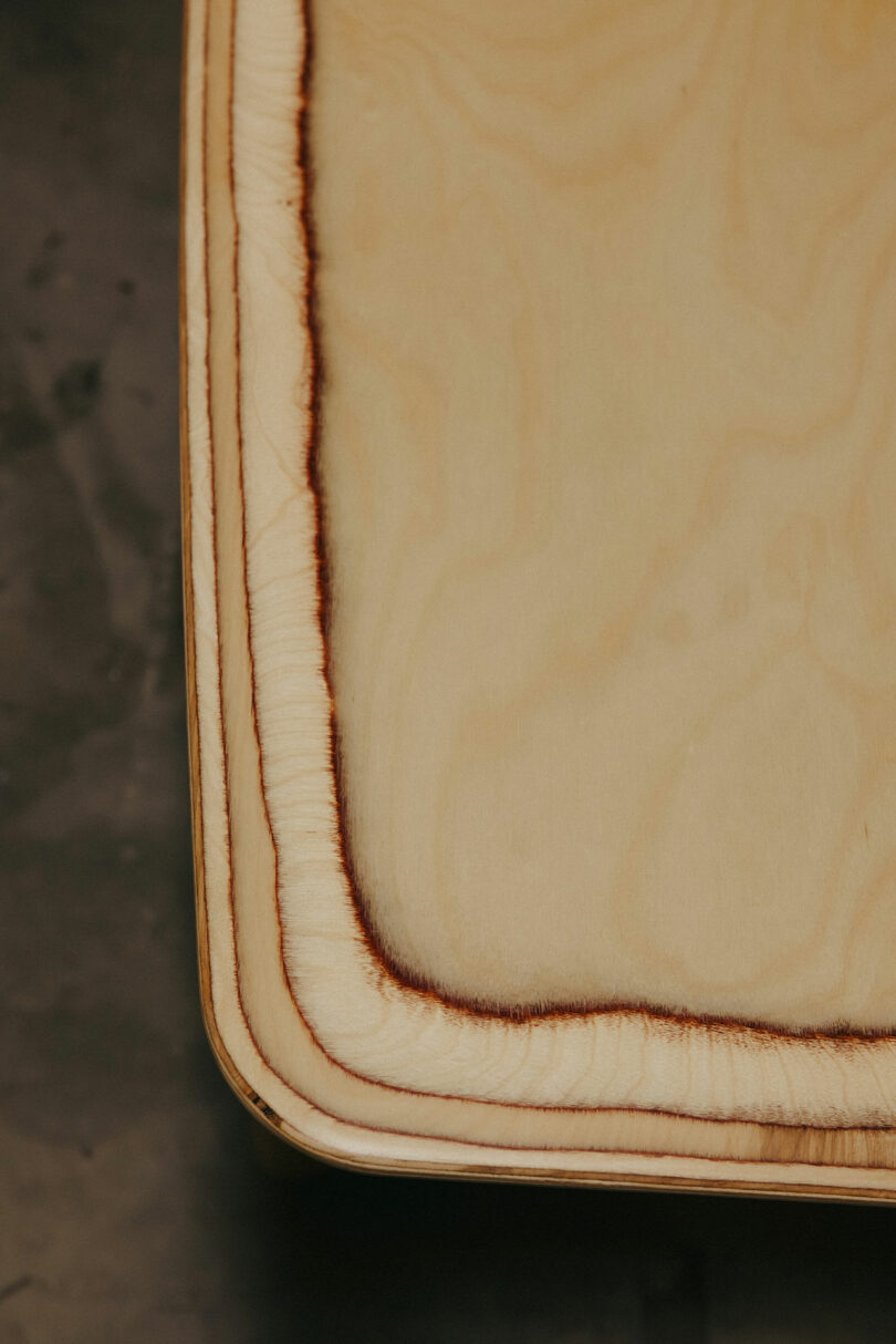 Detail einer leichten Holzoberfläche mit einer von Kelly Wearstler inspirierten abgerundeten Kante mit einer markanten dunklen Brandmarke, die den Rand auf einem neutralen Hintergrund umreißt.