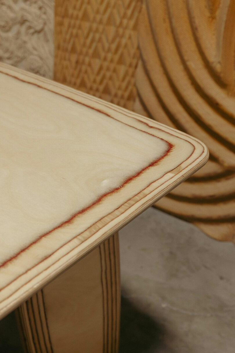 Detail aus Sperrholztisch mit sichtbaren laminierten Kanten, die glatte Oberfläche und natürliche Holzkörner aufweisen. Im Hintergrund erinnern sie geschnitzte hölzerne Objekte moderne Empfindlichkeit von Kelly Wearstler.