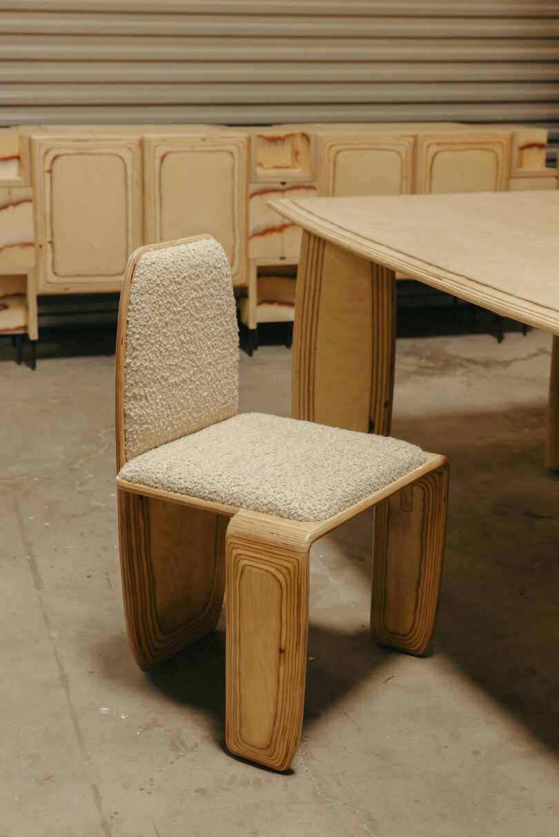 Der Holzstuhl mit gekrümmtem Design und cremefarbenem Kissen, inspiriert von Kelly Wearstler, befindet sich in der Nähe des entsprechenden Holztisches in einer Innenumgebung.