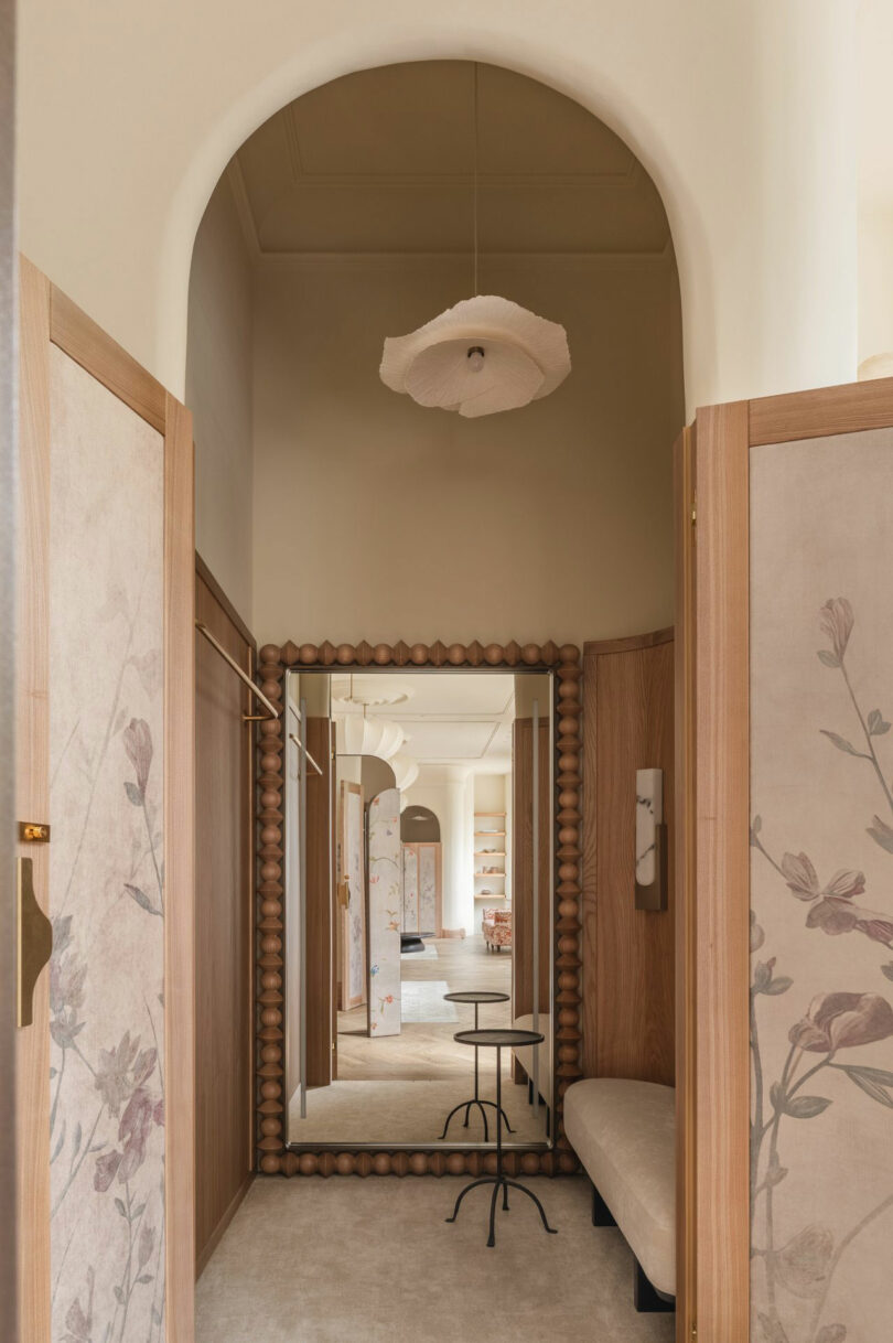 Un corredor con paneles de patrón floral, espejos de madera, un banco amortiguado, una pequeña mesa redonda y un techo escultórico.