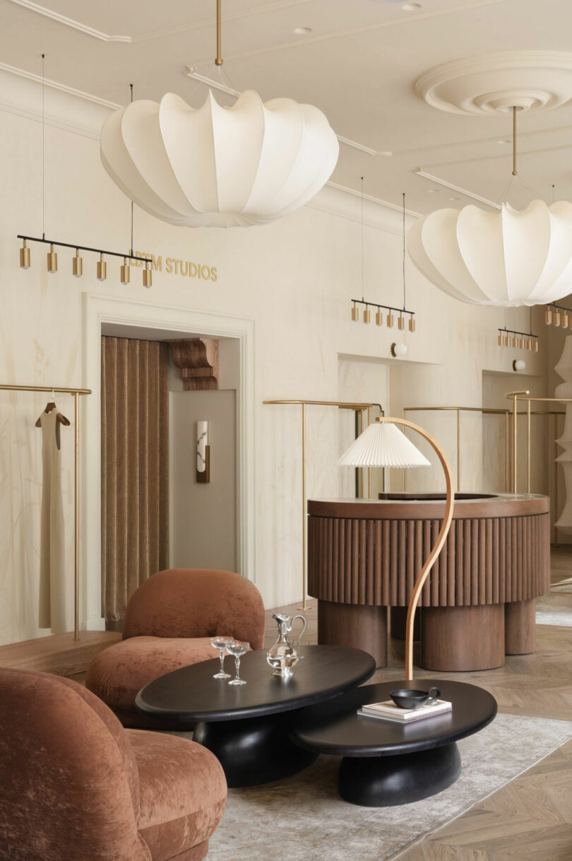 Salón moderno con escritorio de recepción de madera curvo, candelabros de escultura, dos sillas marrones redondas, mesa de anidación negra y decoración con tonos neutros mínimos.