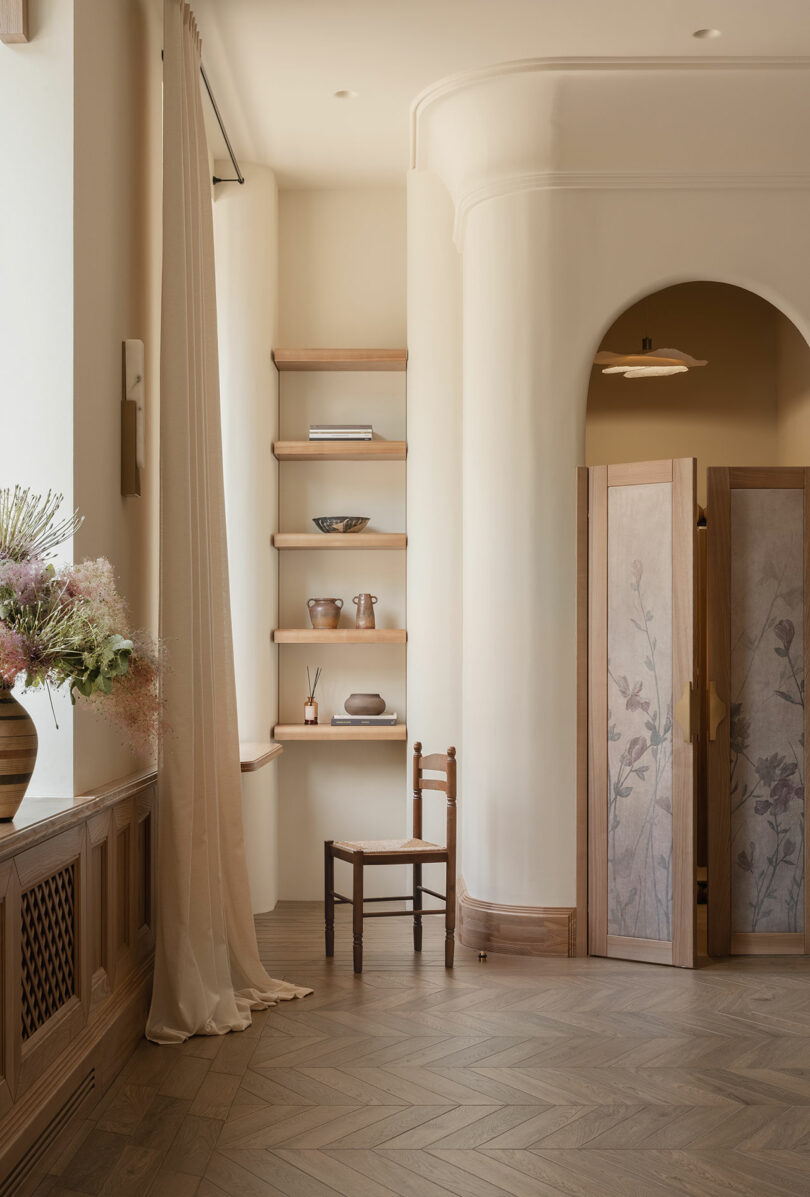 Interior minimalista con sillas de madera, estantes abiertos que exhiben cerámica, puertas arqueadas, pantalla plegable con diseño floral, paredes suaves de beige y jarrón a la izquierda.