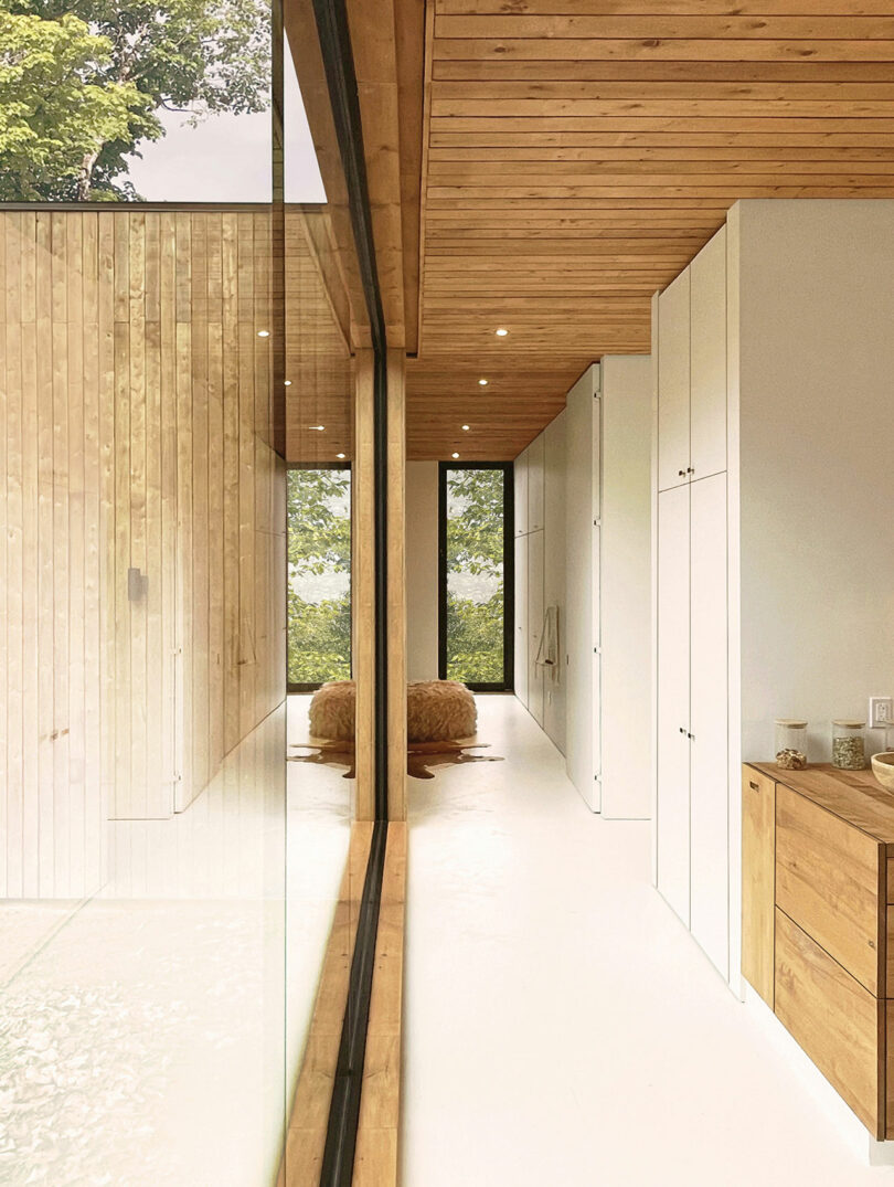 Modern interior hallway with white walls, wooden ceiling, built-in cabinets, and large floor-to-ceiling windows overlooking trees; two brown poufs sit at the end of the hall.