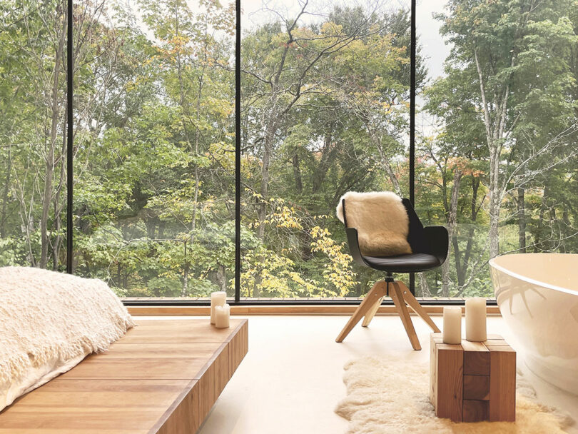 A modern bathroom with large windows overlooking a forest, featuring a wooden platform, black chair with fur throw, white bathtub, and candles.
