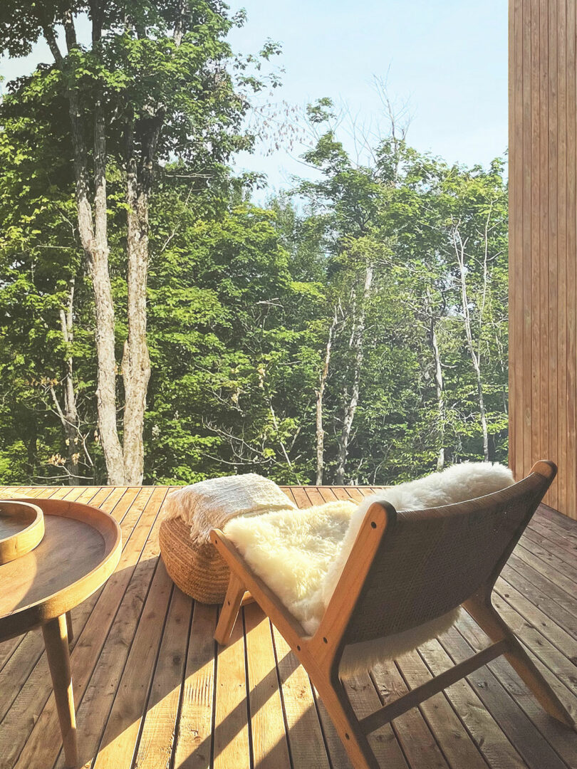 A wooden deck with a lounge chair, sheepskin throw, round ottoman, and side table overlooks a lush, green forest.