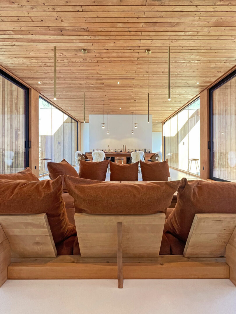 A modern living room with wooden ceiling and walls, large brown cushioned chairs, pendant lights, and a dining area in the background with ample natural light.