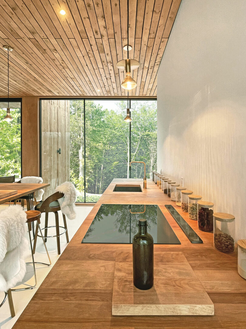 Modern kitchen with wooden countertops, built-in stovetop, glass jars with dry goods, and large windows overlooking a forested area.