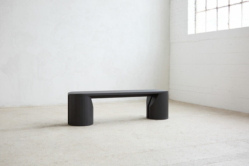 The minimalist dark wooden benches with rounded edges are placed in an empty, light-room room with white walls and a large window.