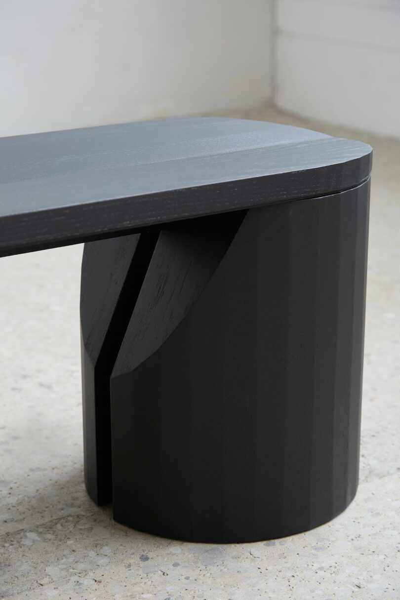 Close-up of a modern black wooden bench with a rounded base and a smooth finish, placed on a light expulsion floor near the white wall.