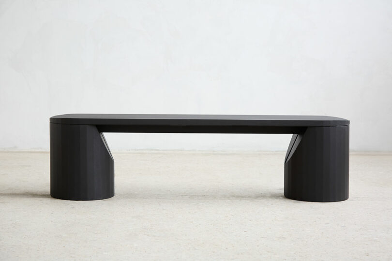 A minimal black bench with a flat seat and two cylindrical legs, placed on a light colored wall and the floor.
