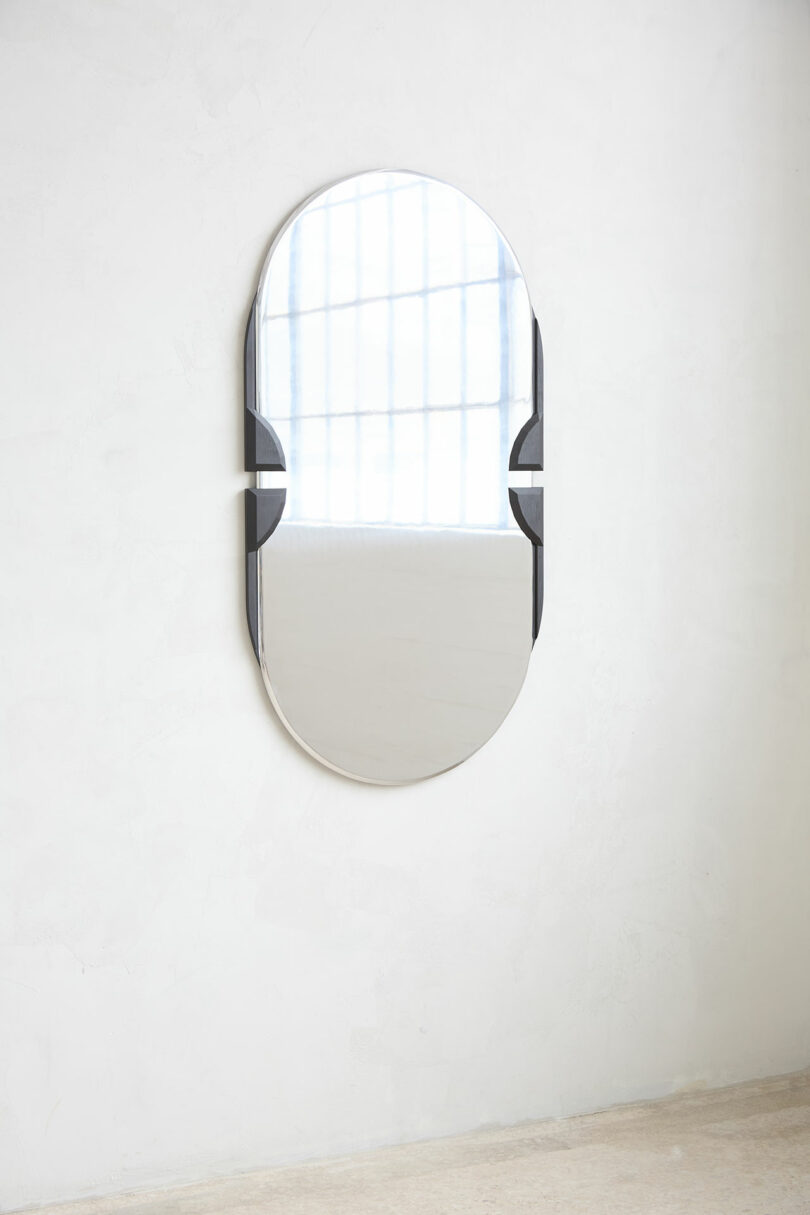 Oval wall mirror with the black frame of the ACCENTS, reflecting a large window with vertical bars, mounts on an ordinary white wall above the light floor.