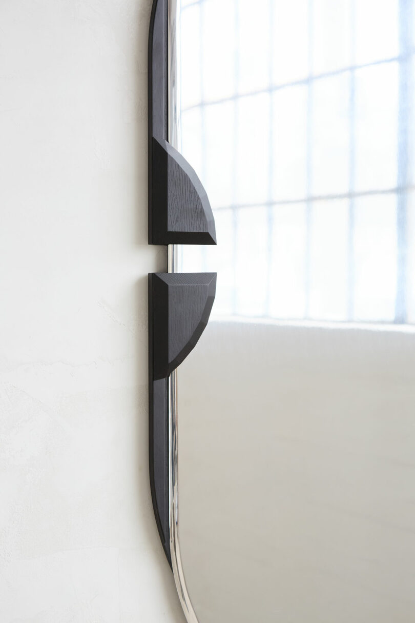 A large rectangular mirror with rounded edges is mounted on a white wall, which contains two geometric black decorative elements on the left.