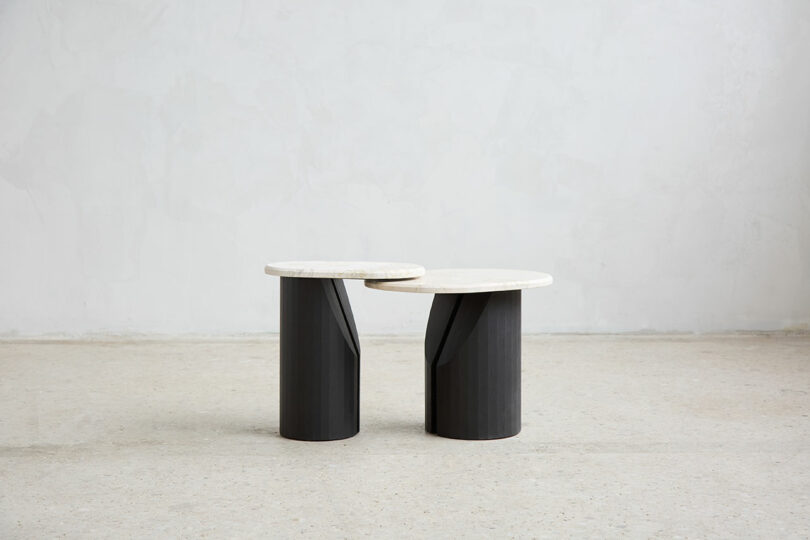 Two modern, round side tables with white tops and black cylindrical bases are placed next to each other on an ordinary, light floor color on the neutral wall.