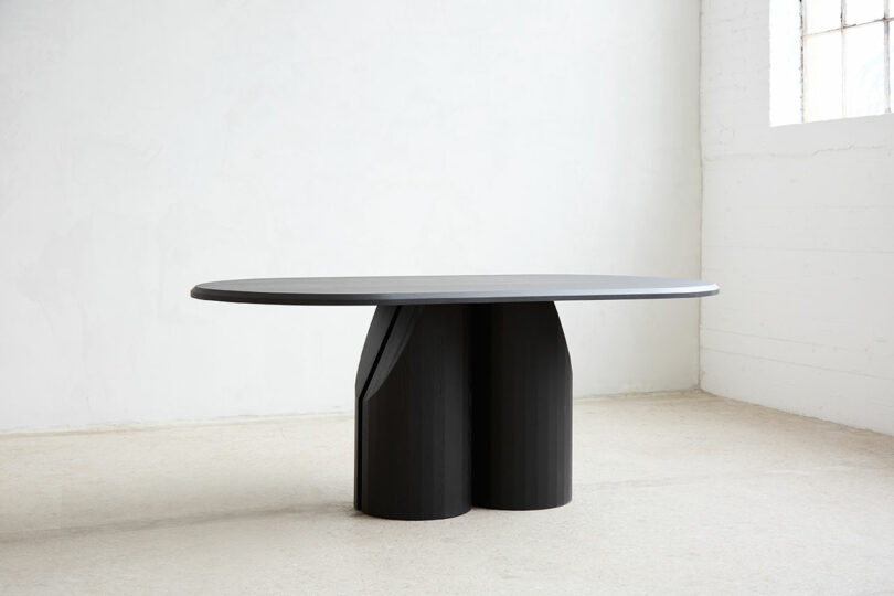 Dark, oval wooden table with a sculptural, rounded base in a minimalist, light room with white walls and window.