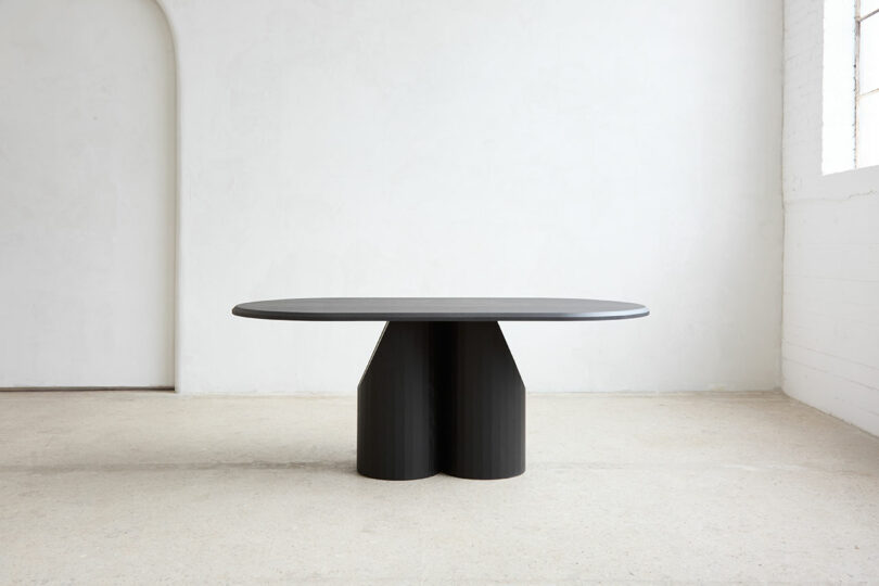 Modern, minimalistic black dining table with rounded rectangular top and two cylindrical stands, placed in a bright, empty room with white walls and light floor.