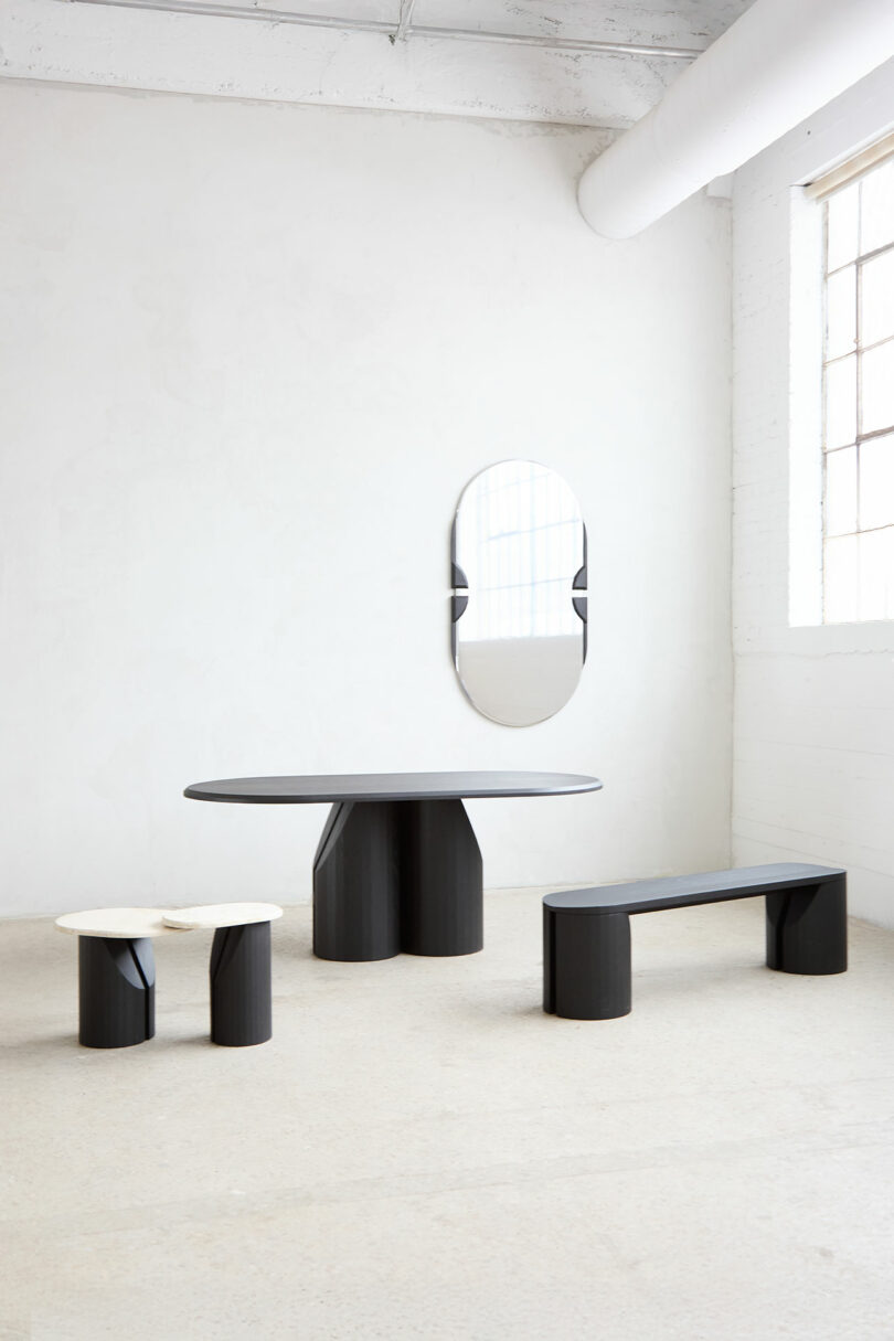 Modern dining set with a black oval table, matching the bench, a small round side table with white tops and an oval mirror on the white wall in the bright room.
