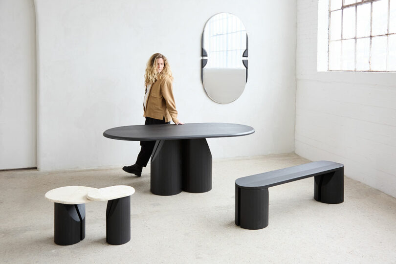 The person stands next to modern black furniture, including an oval table, bench, and a small side table, in a light room with a wall mirror and a large window.
