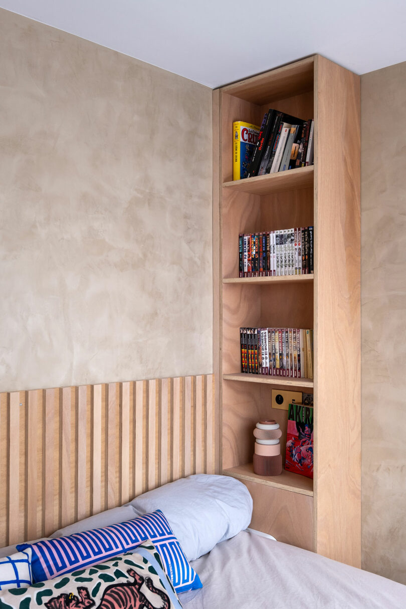 Estantería de madera integrada en una pared de dormitorio sobre la cama, sosteniendo libros, manga y pequeños artículos decorativos cerca de una cabecera de madera de listones.