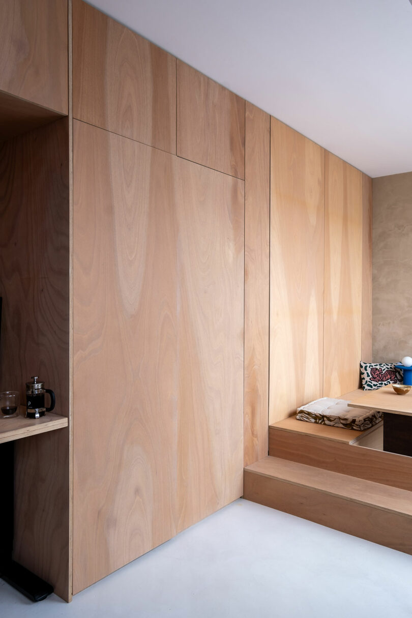 Interior minimalista con paredes de paneles de madera clara, un banco de madera incorporado con cojines y un pequeño estante que sostiene una prensa francesa y vidrio en el lado izquierdo.