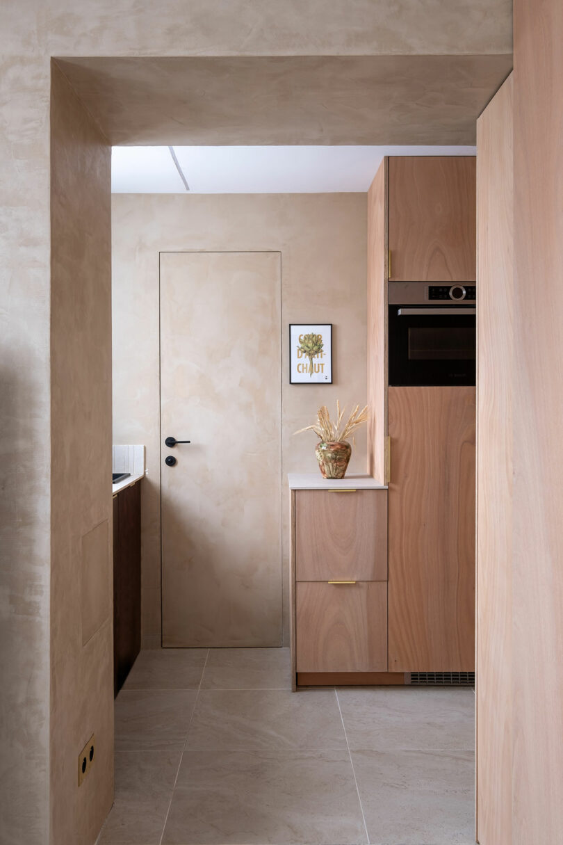 Cocina minimalista con gabinetes de madera ligera, paredes beige, puerta cerrada, electrodomésticos incorporados y una pequeña obra de arte enmarcada sobre un jarrón en la encimera.
