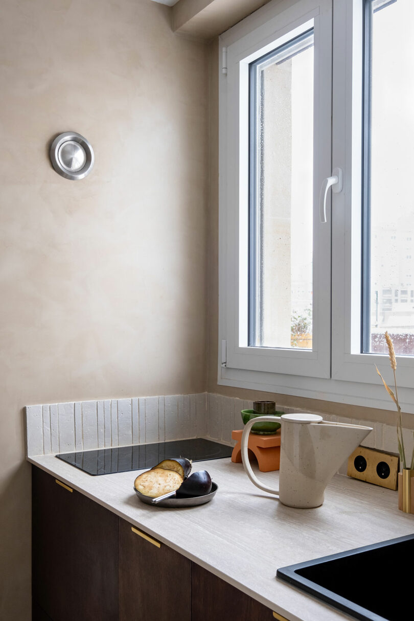 Una encimera de cocina minimalista con una tabla de cortar que sostiene el pan en rodajas y una berenjena, una jarra beige, dos pequeñas plantas en macetas y una estufa debajo de una ventana grande.