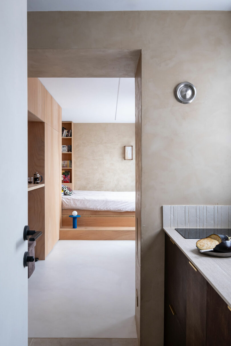 Vea desde una cocina a un dormitorio minimalista con muebles de madera ligera, estantes empotrados y una cama cuidadosamente hecha.