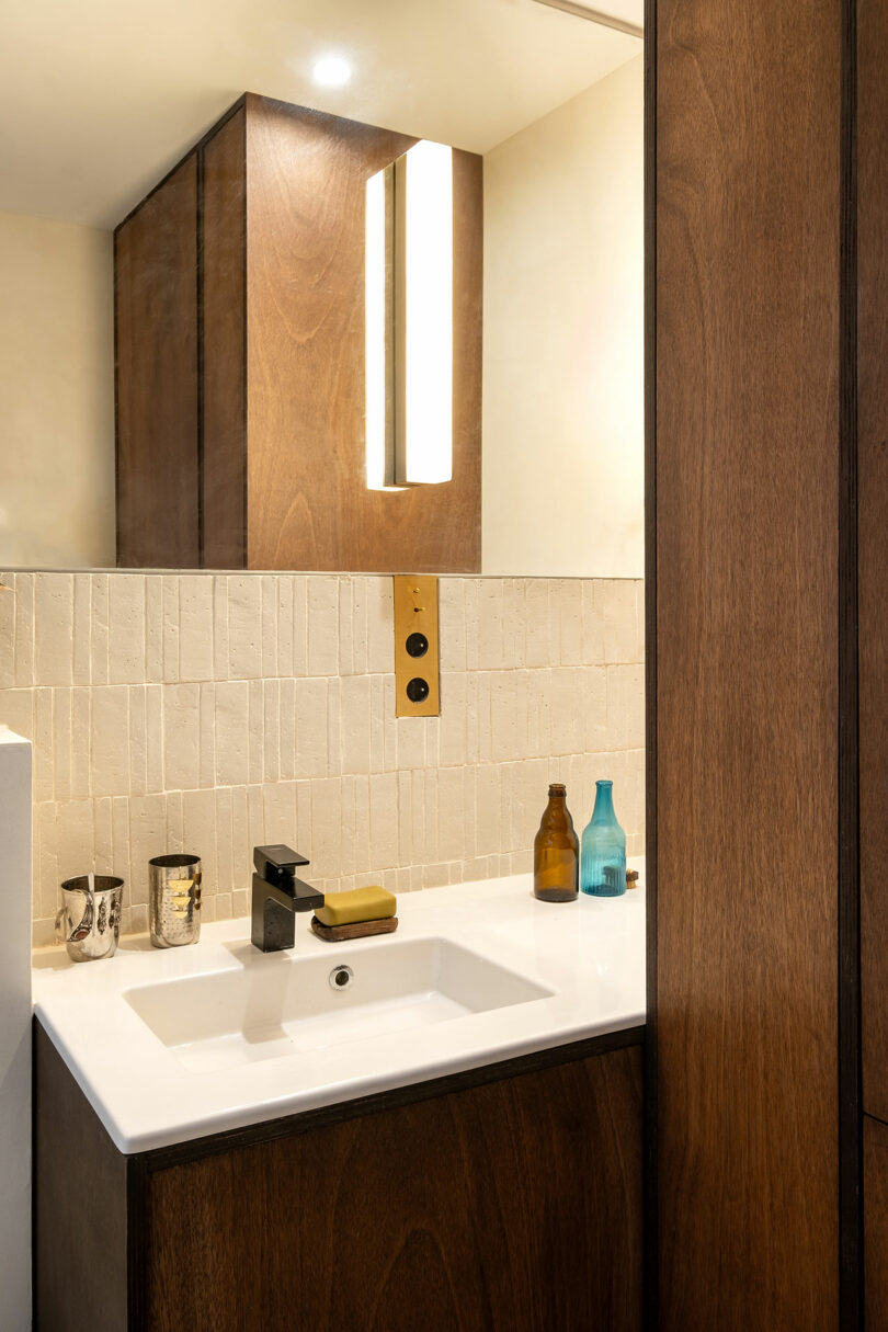 Moderno lavabo de baño con una cuenca blanca rectangular, grifo negro, jabón y botellas en el mostrador. La luz vertical de la pared y los gabinetes de madera son visibles en el fondo.