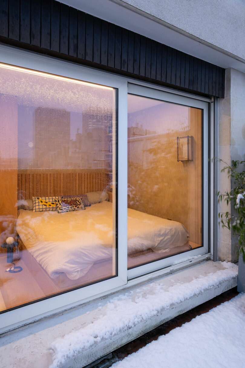Una acogedora habitación con una cama doble y almohadas estampadas se ve a través de grandes puertas de vidrio, con nieve que cubre el suelo y la repisa de las ventanas afuera.