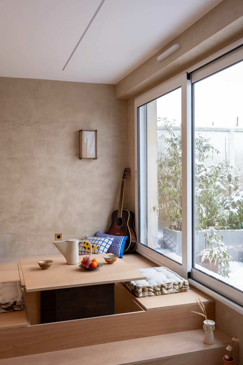 Habitación minimalista con una mesa de madera hundida, tazones, frutas, cojines, una guitarra y una gran ventana que muestra una escena nevada al aire libre con plantas.