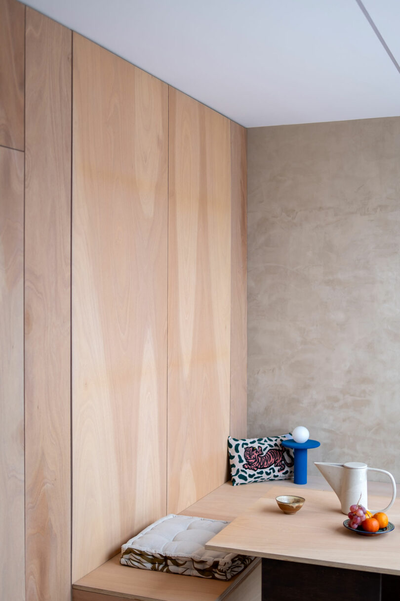 Rincón de comedor minimalista con paneles de madera clara, una pequeña mesa con una jarra blanca, tazón, fruta y una lámpara azul, además de cojines estampados en los asientos de banco.