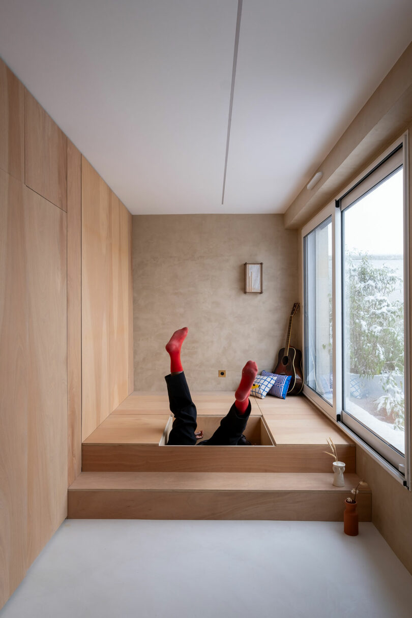 La persona con calcetines rojos se encuentra en la espalda con las piernas en una habitación de madera minimalista, al lado de una ventana, una guitarra y una mesa baja, con luz natural.