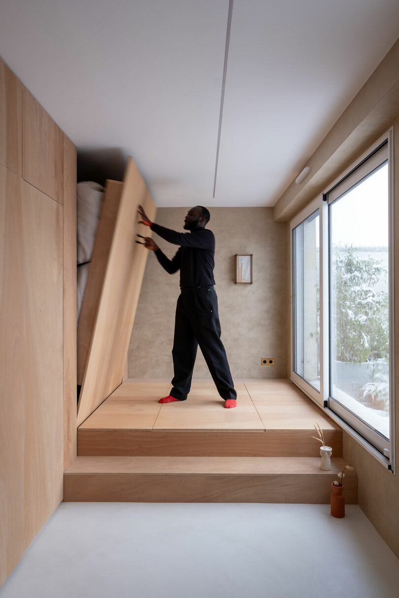 Una persona levanta una cama plegable integrada en una plataforma de madera en una habitación minimalista y moderna con luz natural.