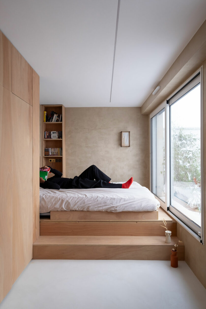 Persona acostada en una cama elevada que lee un libro en un dormitorio minimalista con paneles de madera ligera, estantes incorporados, ventana grande y paredes de color neutral.