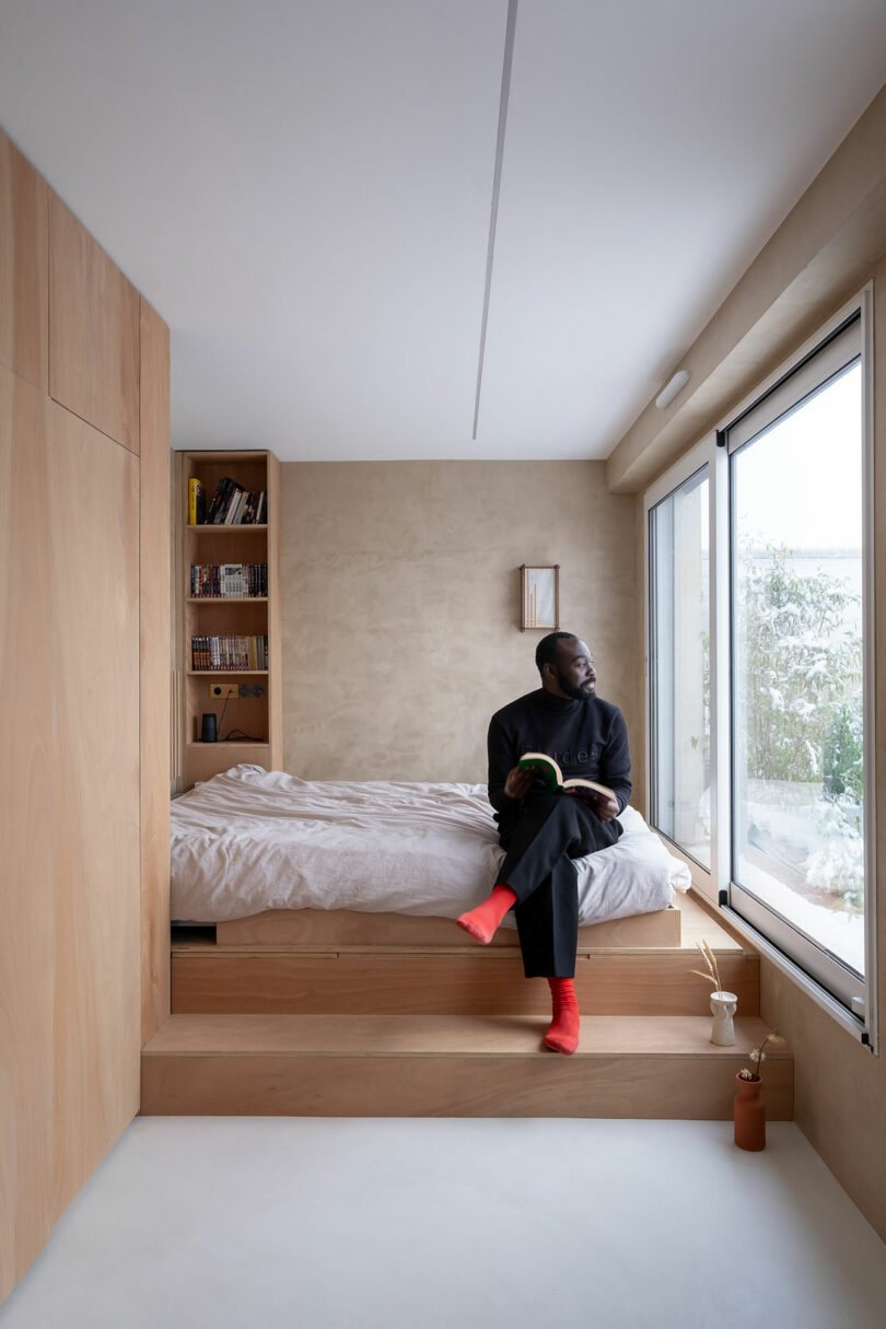 Un hombre con calcetines rojos se encuentra en el borde de una cama en una habitación minimalista con acentos de madera, sosteniendo un libro y mirando por una gran ventana.