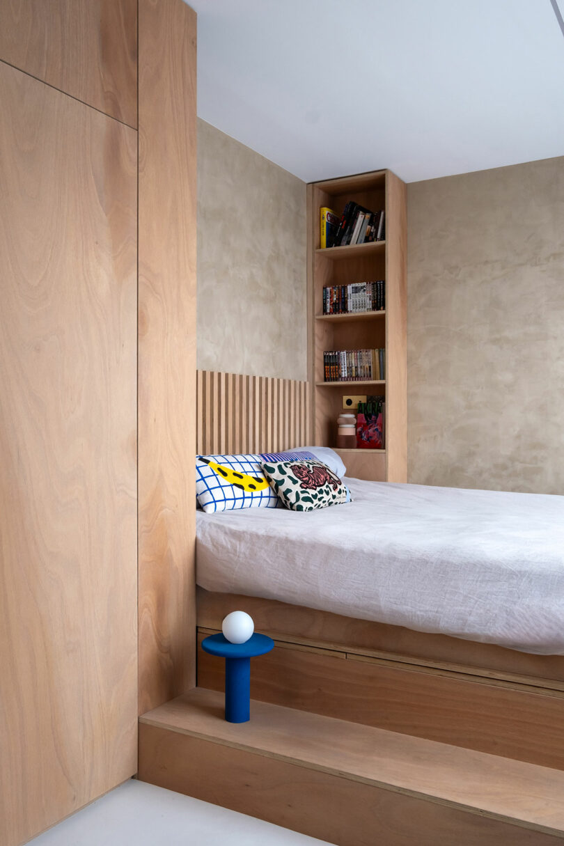 Un dormitorio moderno con paneles de madera clara, una estantería incorporada, una cama con cojines estampados y una pequeña mesita de noche azul con una lámpara esférica.