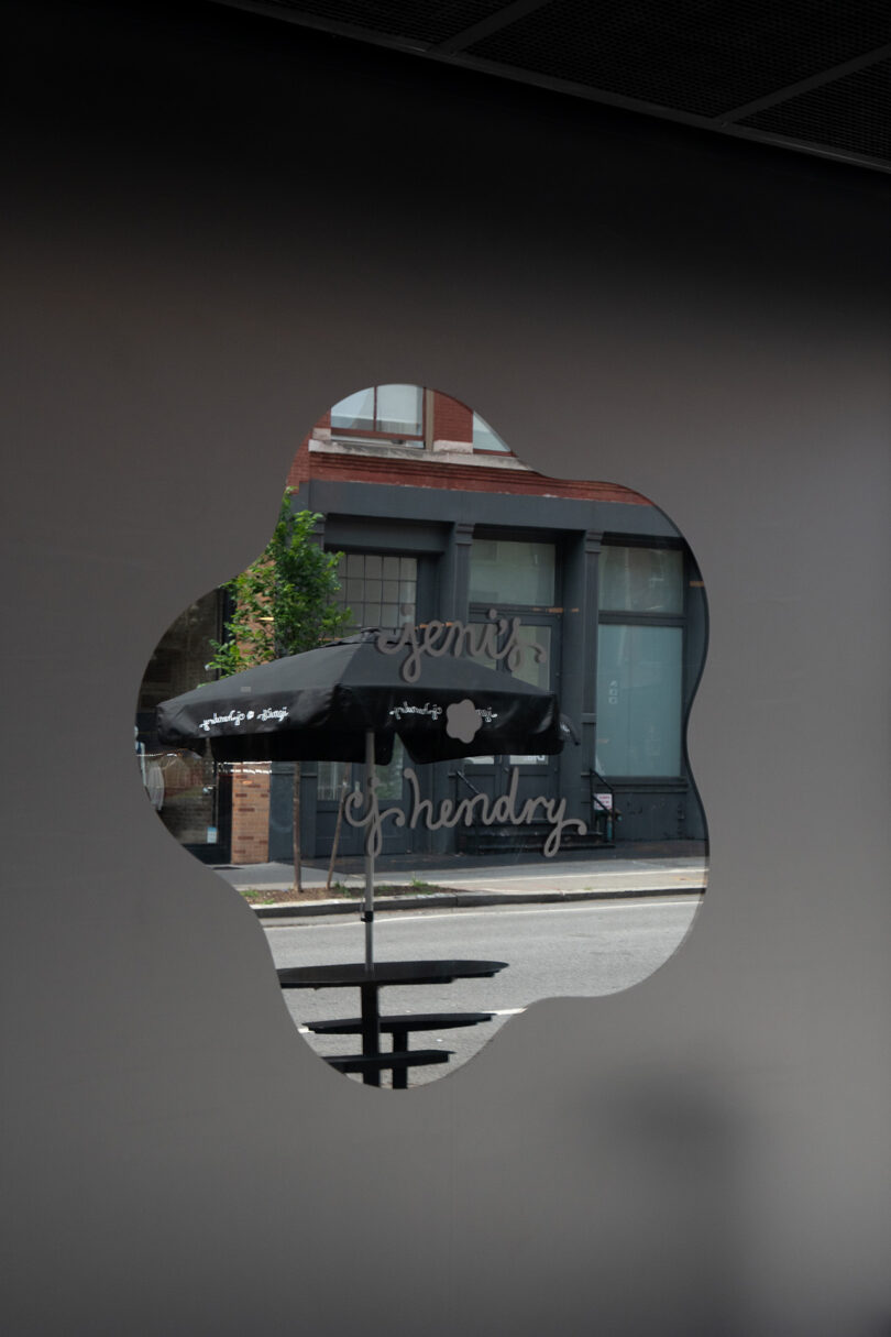 A window with an OPAQUE, cloud-shaped cutout reveals a street view, featuring a building and a black umbrella with "Jeni's Splendid" visible.