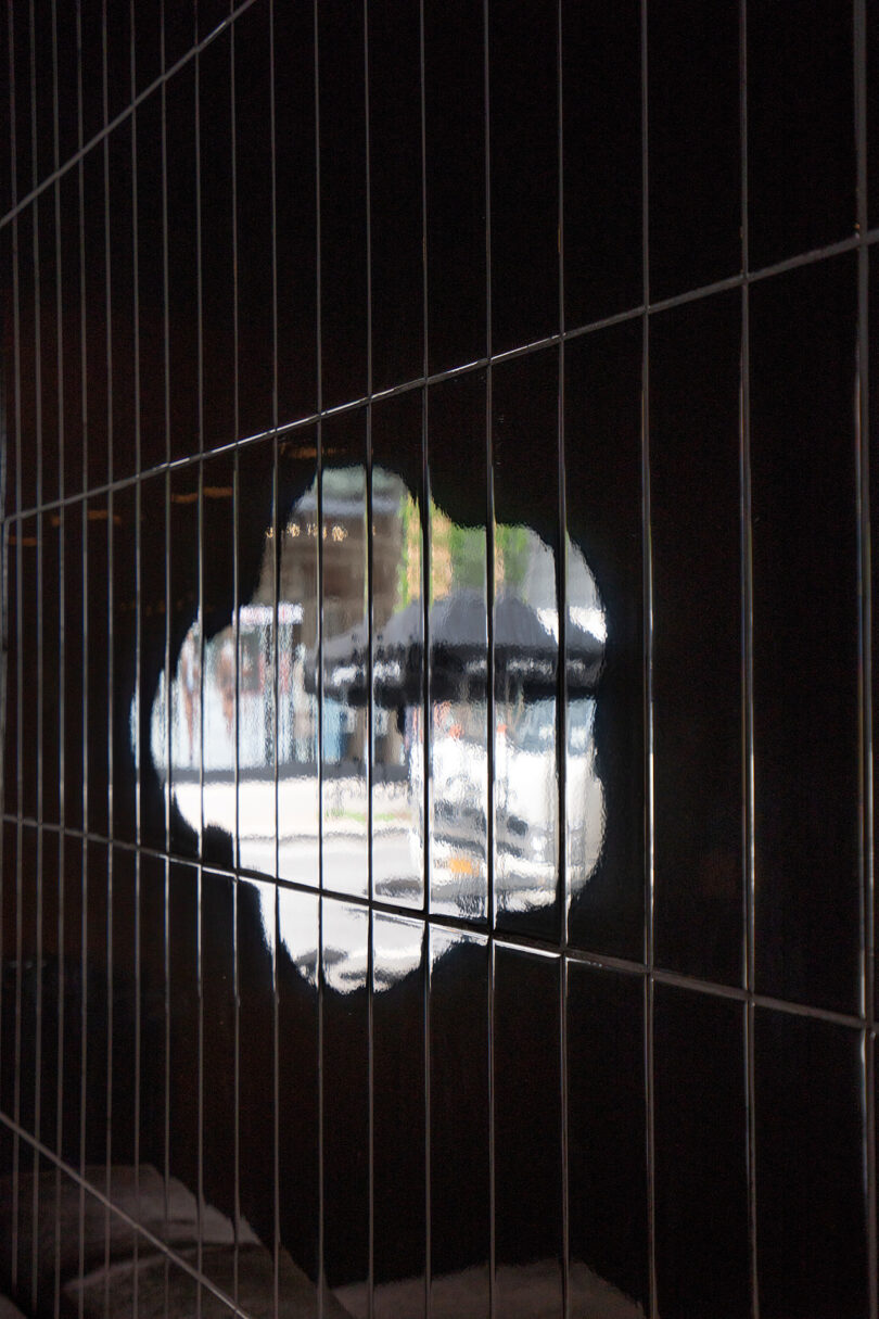 A reflective tiled wall features an irregularly shaped clear spot that reveals a blurred street scene with people and outdoor umbrellas outside.