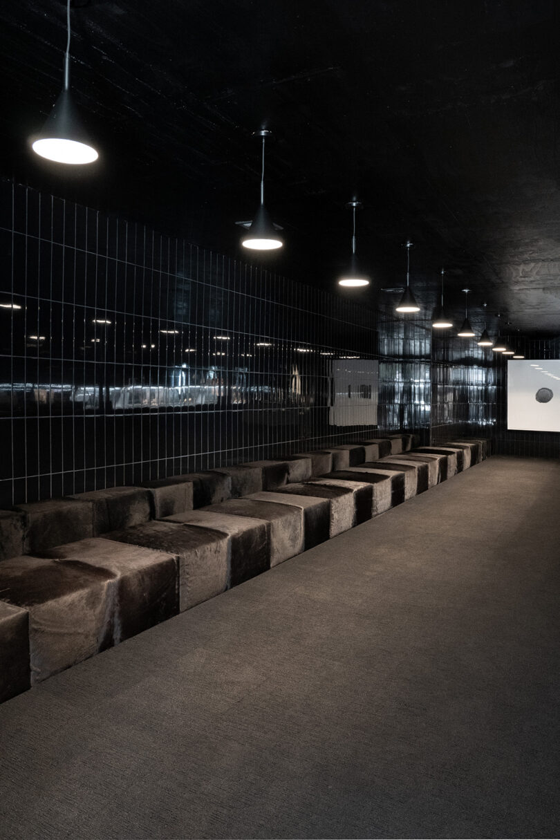 A modern interior with black tiled walls low cushioned seating along one side, pendant lights overhead, and a grey carpeted floor.