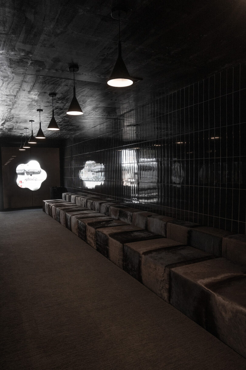 A dimly lit hallway with black tiled walls, pendant lights, and a long row of cushioned seating along one side; light enters from patterned windows at the end.
