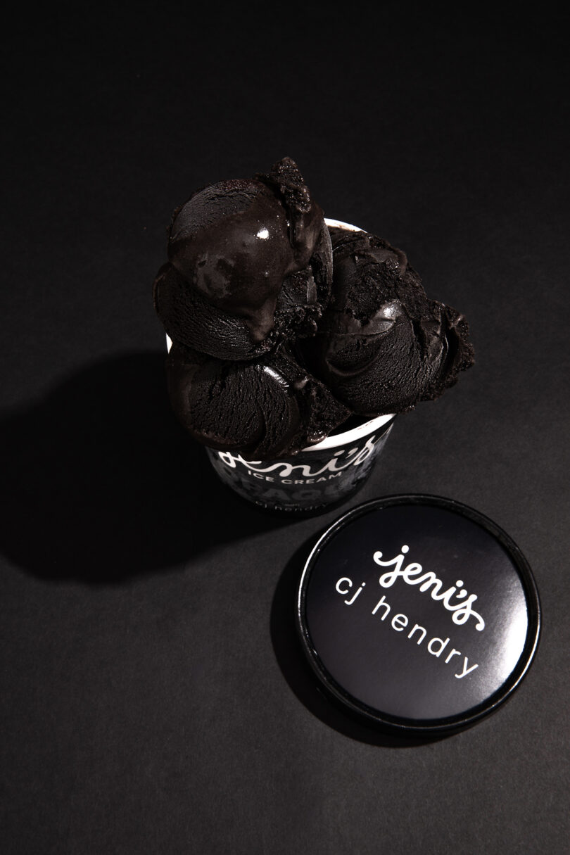 A cup of dark black ice cream from Jeni's with a branded lid labeled "jeni's cj hendry" placed beside it on a black background.